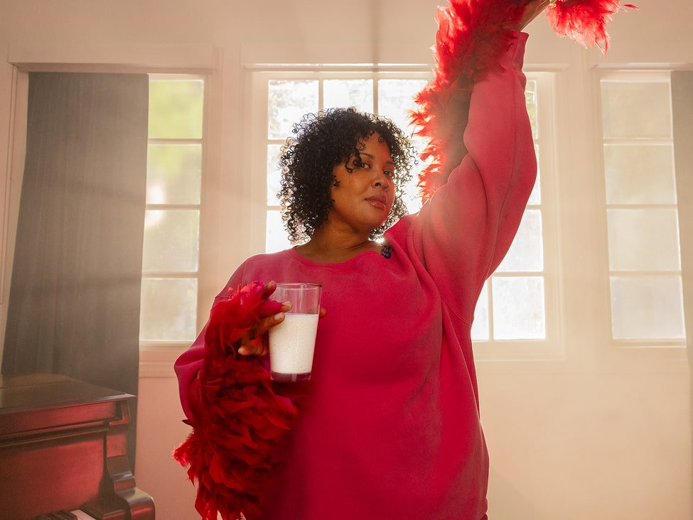 A woman in a bright pink outfit holds a glass of milk and raises a red feather boa in the air, standing confidently in front of sunlit windows.