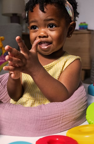 A smiling toddler in a yellow dress and pastel headband claps her hands while playing with colorful toys in a cozy living room.