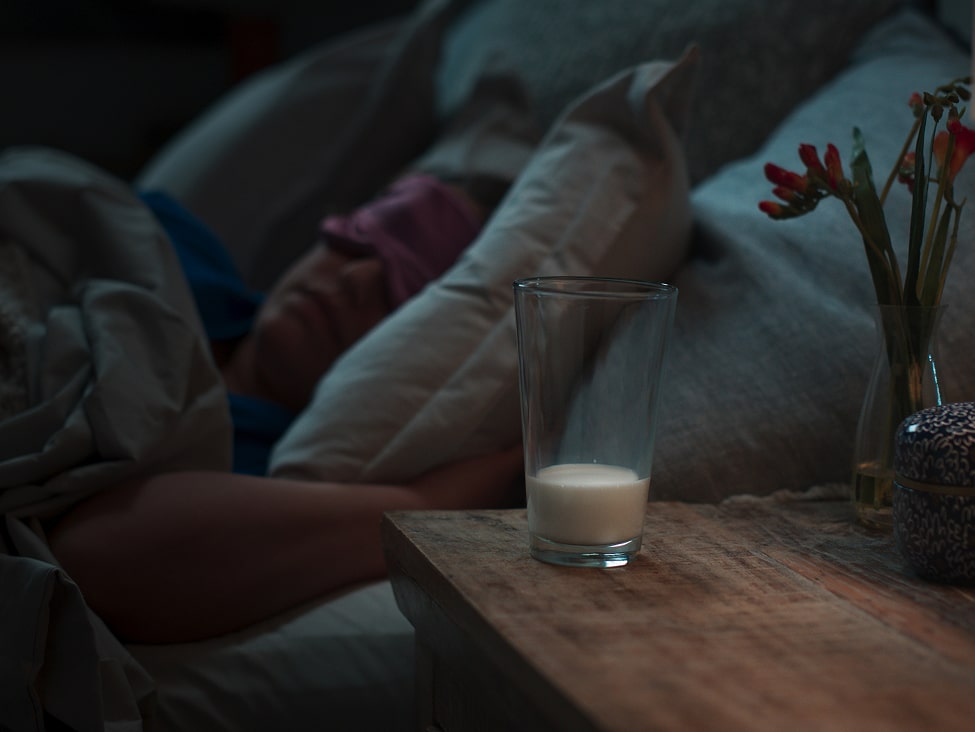 A glass of milk on a wooden bedside table with a sleeping person in the background wearing a pink sleep mask, surrounded by pillows and dim lighting.