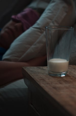 A glass of milk on a wooden bedside table with a sleeping person in the background wearing a pink sleep mask, surrounded by pillows and dim lighting.